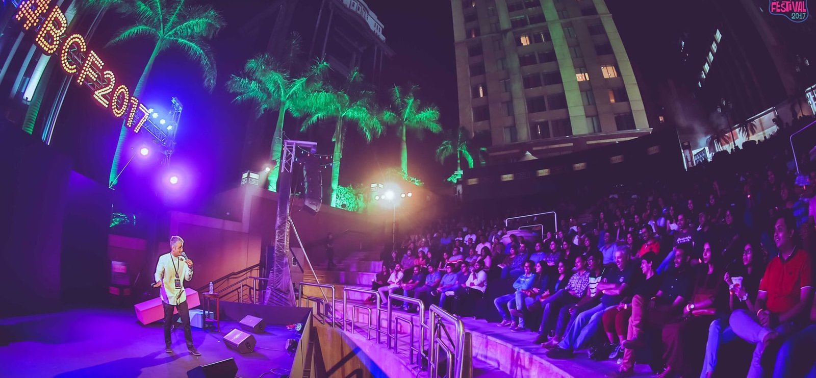 Comedian performing on an outdoor festival stage to a large amphitheater crowd at night