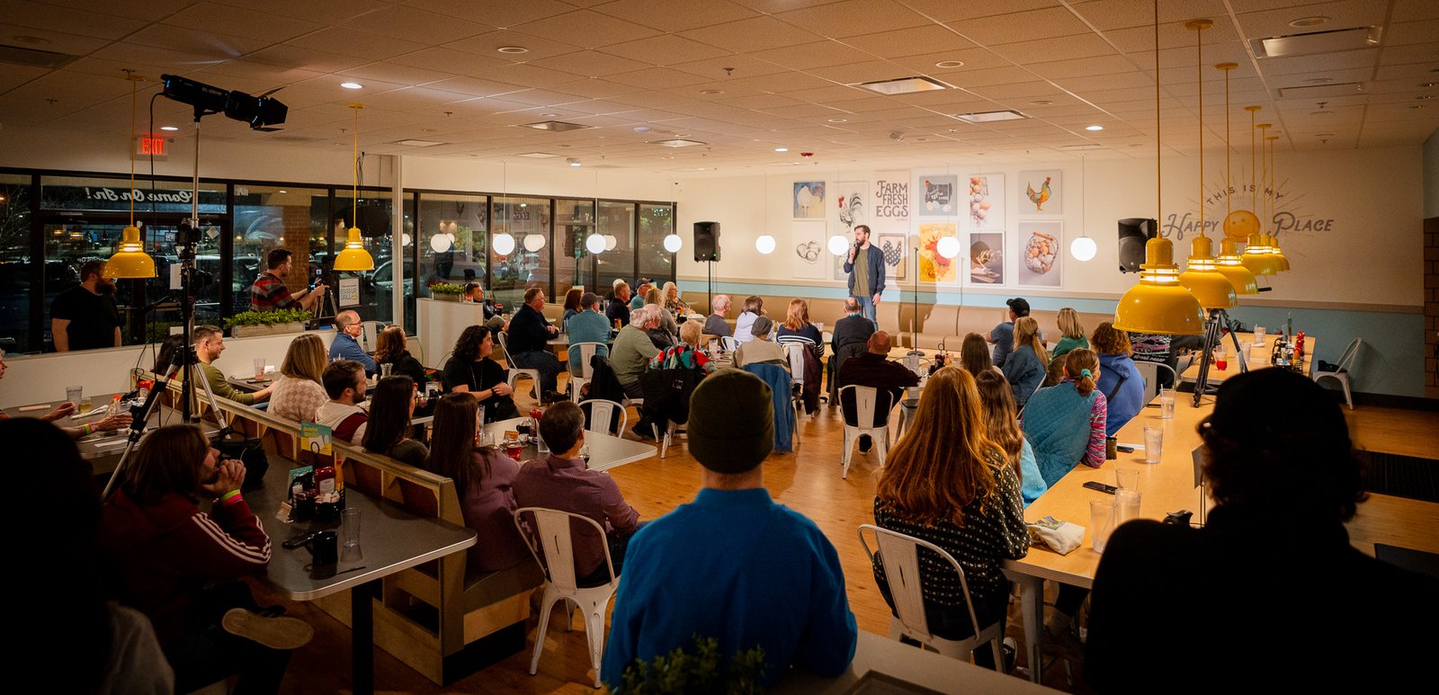 Comedian performing to a full casual dining room, seen from the back of the seated crowd