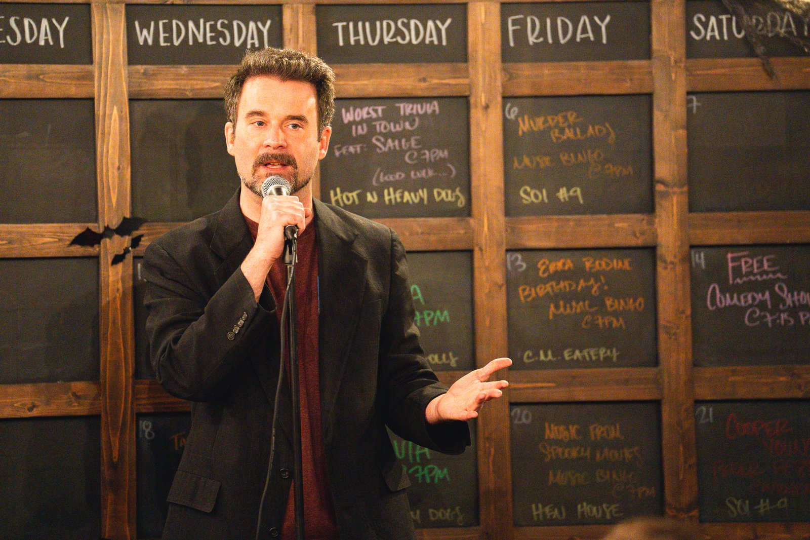 Comedian in a dark blazer working a handheld microphone in front of a wall of chalkboard show listings