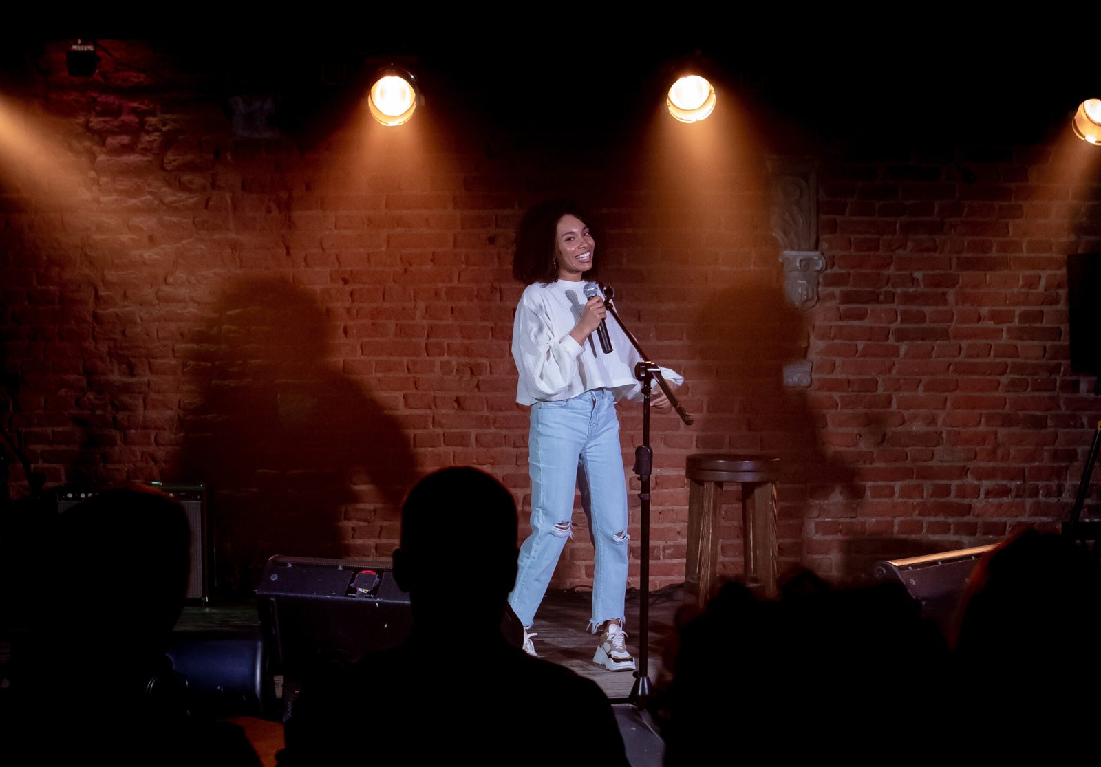 Comedian smiling at a microphone stand on a brick-walled club stage under warm spotlights
