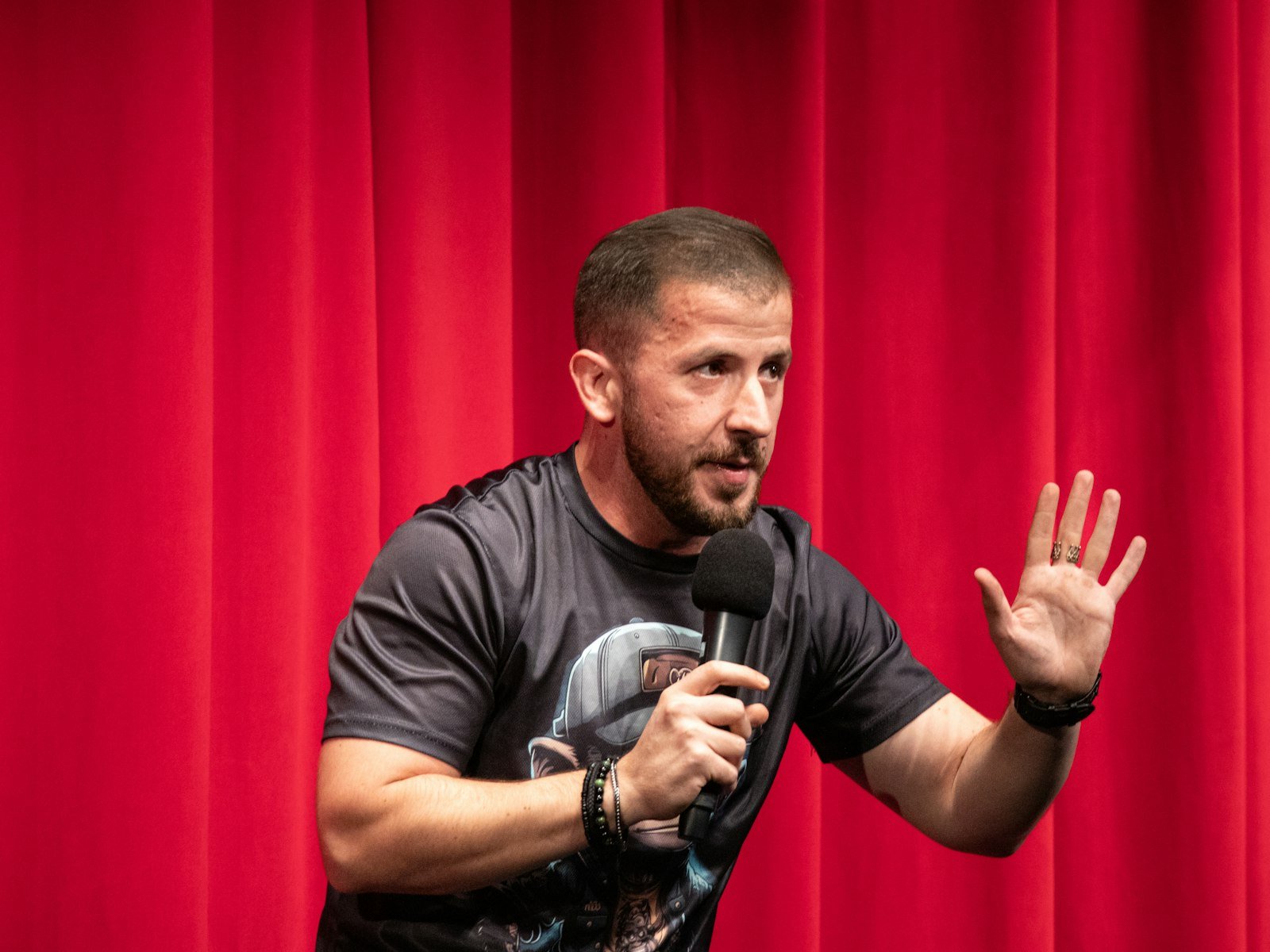 Comedian in a T-shirt gesturing mid-joke in front of a red curtain
