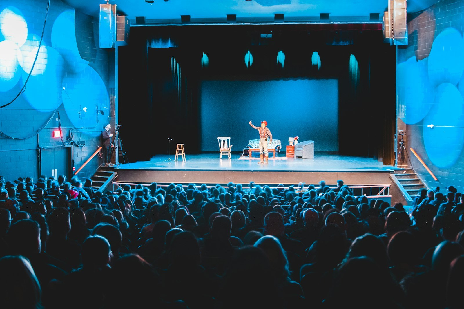 Audience in a large theatre facing a blue-lit stage with two chairs set for a show