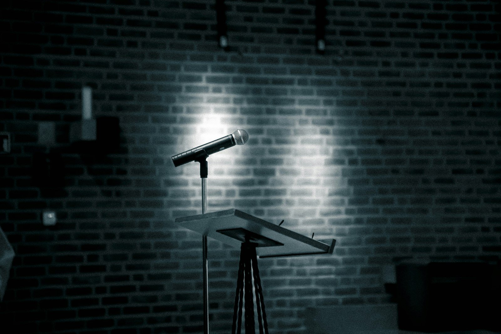 Microphone on a stand in front of a bare brick wall, lit by a single spotlight above an empty lectern
