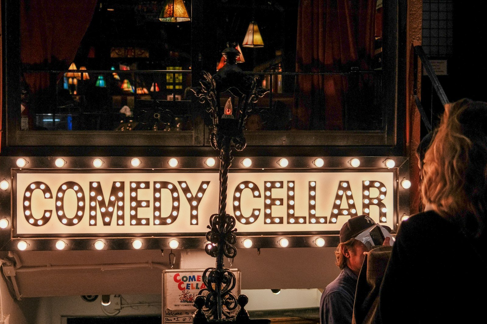 Lit Comedy Cellar marquee sign over a doorway with people waiting outside on the sidewalk at night