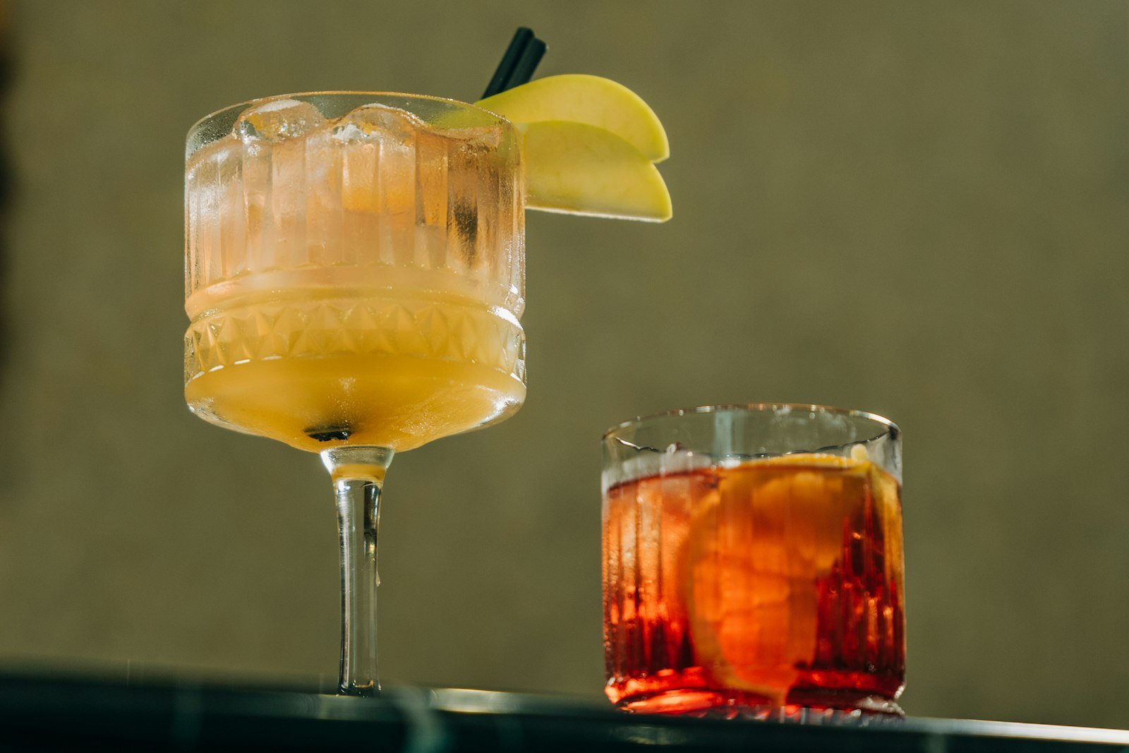 Two cocktails on a bar top, one with apple slices on the rim and one served red over a large ice cube