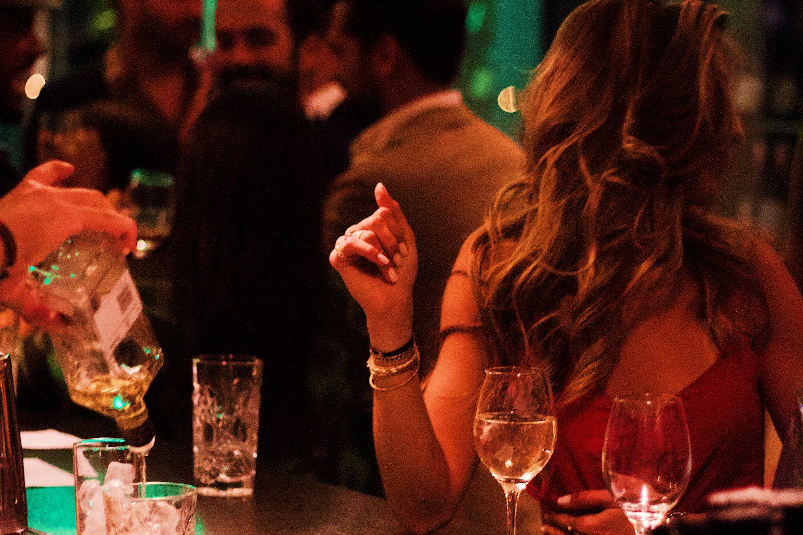Busy bar counter at night with wine glasses in the foreground and a bartender pouring a drink