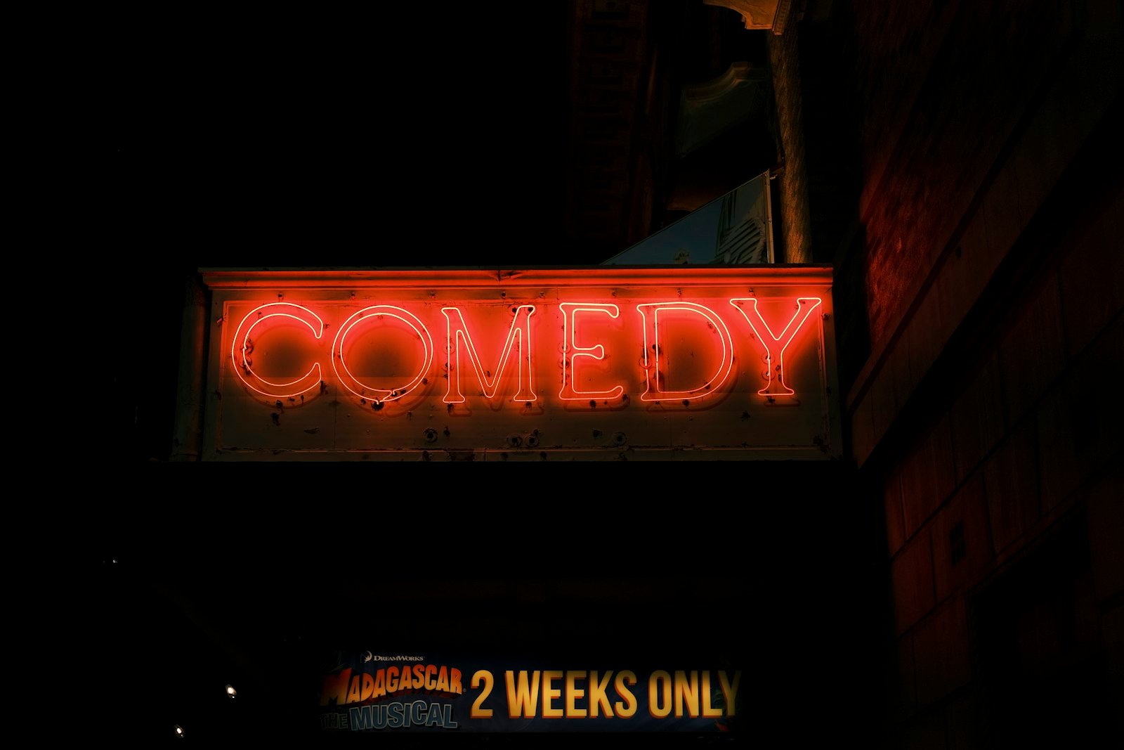 Red neon sign reading COMEDY glowing over a dark theatre entrance at night