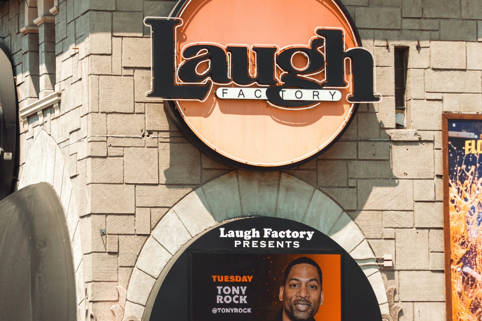 Round Laugh Factory neon sign on a stone wall above a lit marquee board advertising a Tuesday headliner