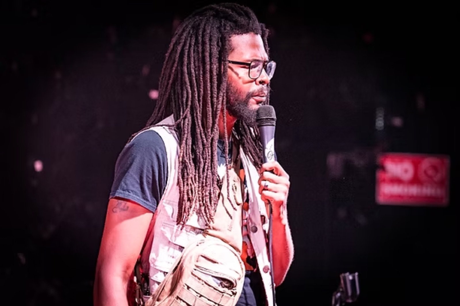 Stand-up comedian with long locs performing on a dark club stage with a handheld microphone