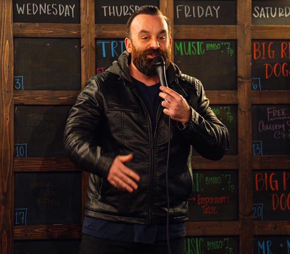 Bearded comedian in a leather jacket performing in front of a chalkboard wall listing the bar's weekly events
