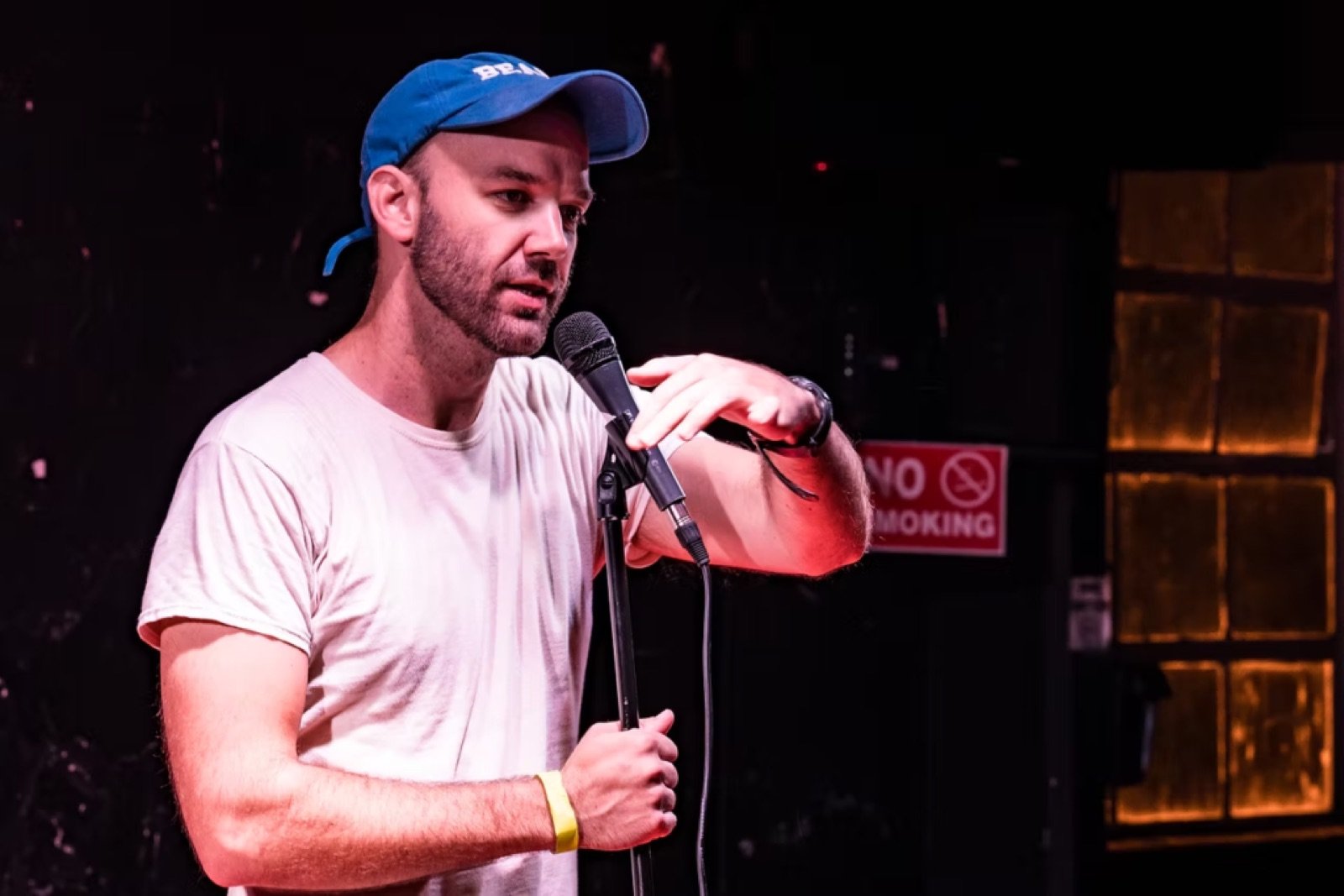 Stand-up comedian in a blue cap performing at a microphone stand in a dark club