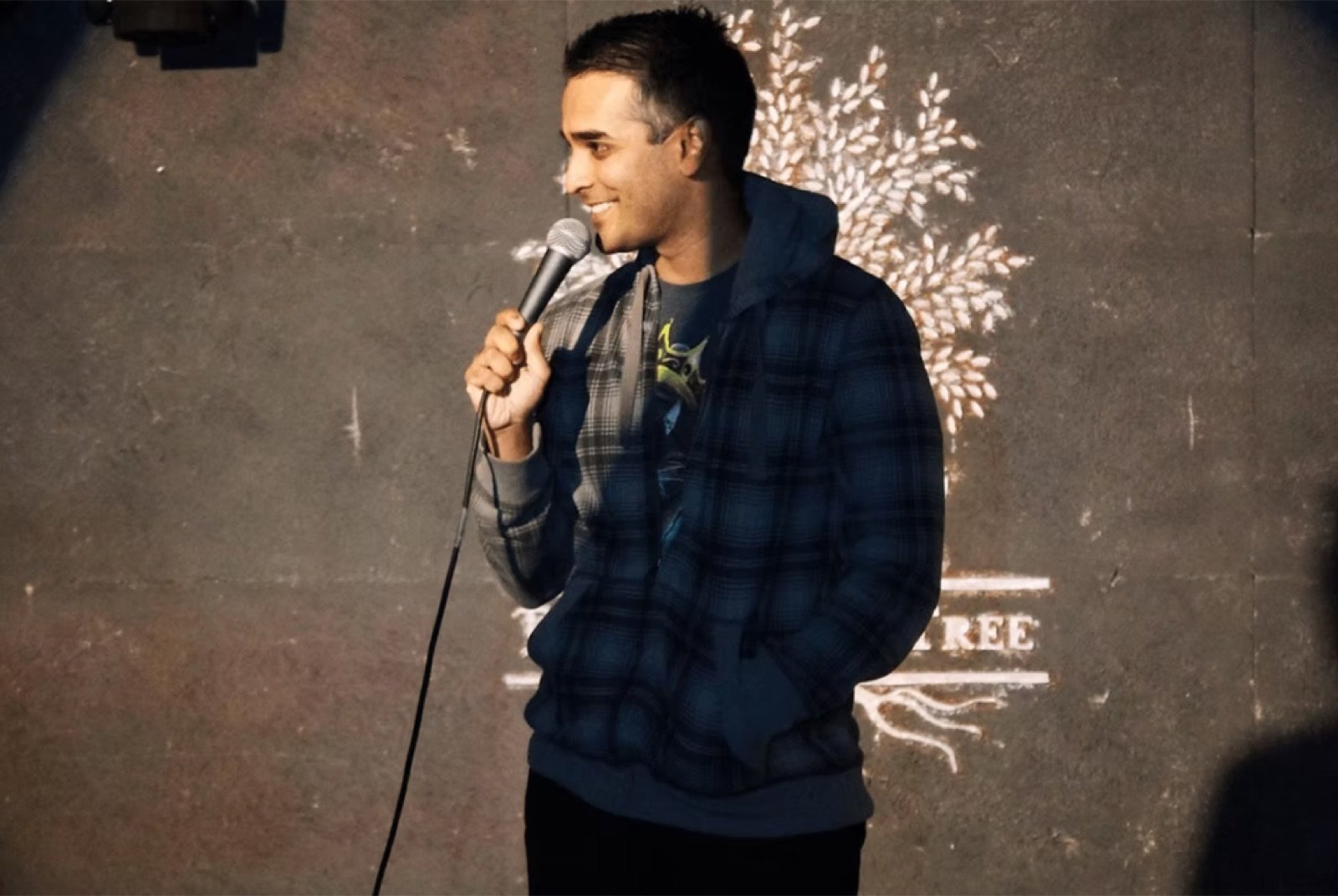 Comedian smiling with a handheld microphone in front of a painted stage wall