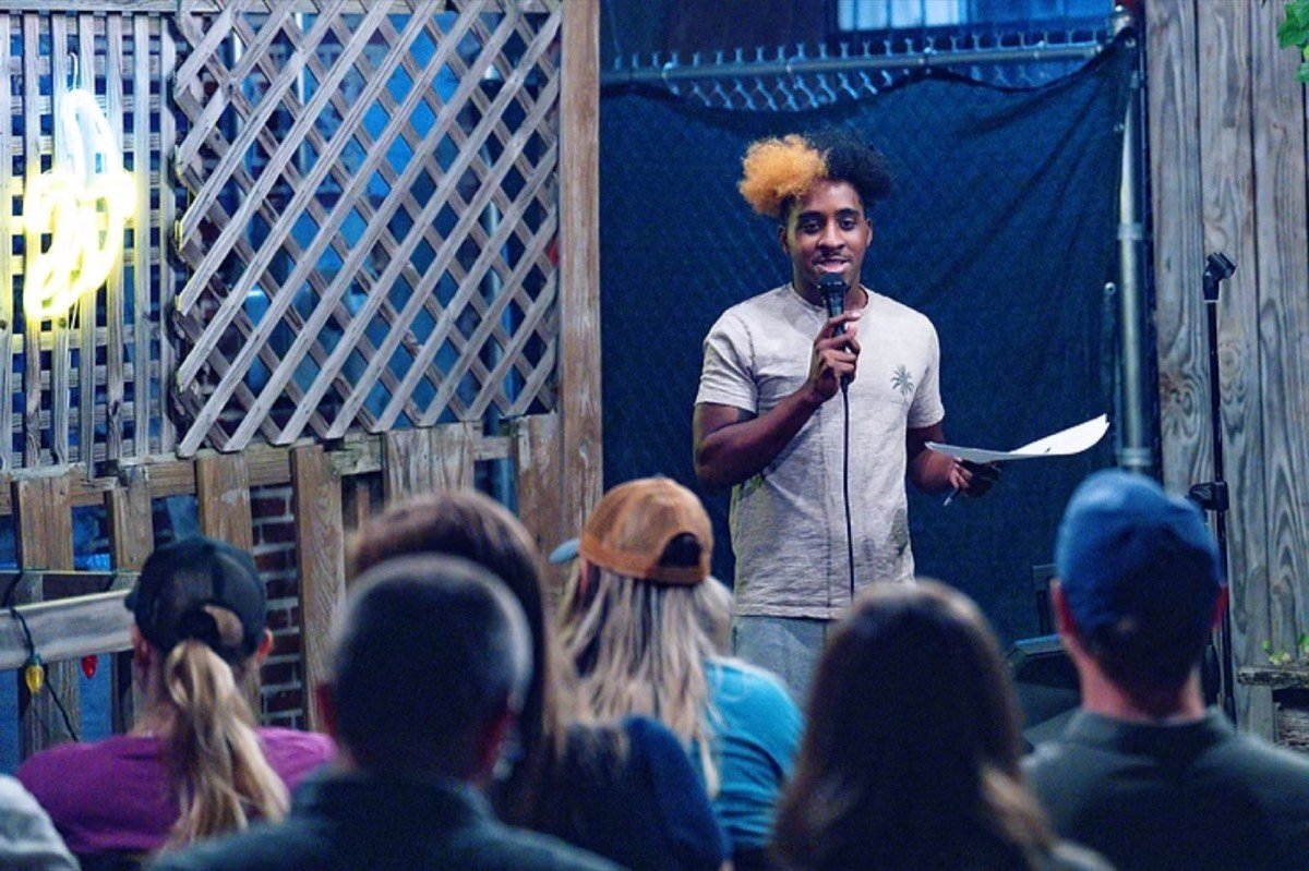 Comedian performing on a small outdoor patio stage in front of a seated audience