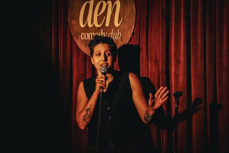Comic performing on stage at The Den Comedy Club in Houston, red curtain and the club's round wooden sign behind her